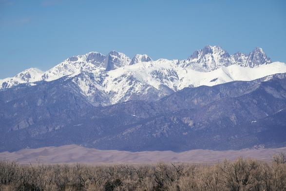 Snow-capped mountains with rugged peaks, sand dunes in the midground, and bare trees in the foreground under a clear blue sky.