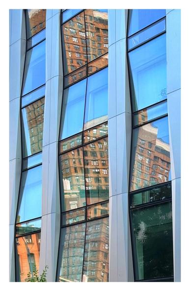 Closeup of the facade of the Luminaire, a luxury condo in the NoMad (north of Madison Square Park) neighborhood of Manhattan, NYC. The photo is taken from street level and at an angle such that the facade faces toward upper left. Beveled pilasters frame multiple levels of windowpanes which are alternately angled upward and downward. This breaks up the reflection of a red brick high-rise apartment building across the street, in a sort of checkerboard effect: some panels reflect portions of the high-rise (with its own facade of many windows), while others reflect only a clear blue sky.