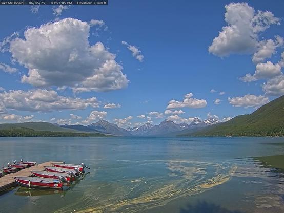A blue sky filled with puffy white clouds hangs over a group of red, silver, grey, and white boats lining both sides of a dock. The lake is Lake McDonald in Glacier National Park. The image is of interest for the swirl of yellow in the foreground. It is likely that this swirl of yellow is pollen, set in motion and sculpted by the wind. Snow-capped mountains and green tree-covered slopes appear in the distance. Thanks to the National Park Service and the Glacier National Park Conservancy for providing these beautiful views.