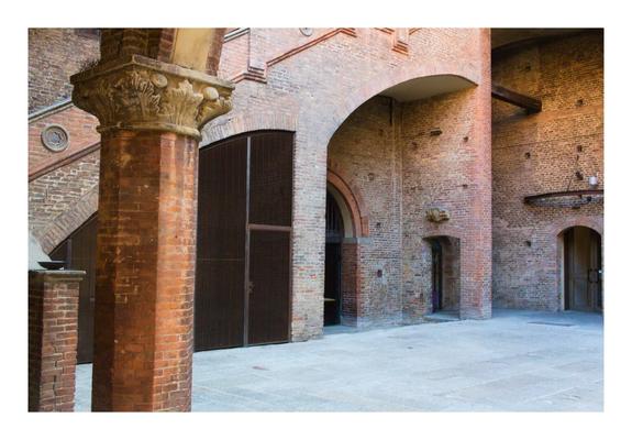 This photo shows the inner courtyard of a historic brick building, Palazzo Re Enzo in Bologna, Italy. The walls are made of reddish-brown bricks with arched doorways and windows. A thick, round column stands in the foreground, topped with a detailed stone capital decorated with carved leaves. The ground is paved with large stone tiles. Wooden doors and iron fixtures are visible throughout. A staircase with a brick railing rises in the background, leading to an upper level. The scene is softly lit by natural light, creating gentle shadows that highlight the architectural textures and the aged, rustic character of the space.
