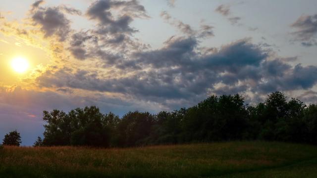 Evening photo of a meadow and a treeline. There are some scattered clouds in the sky lit orange/yellow by the setting sun.