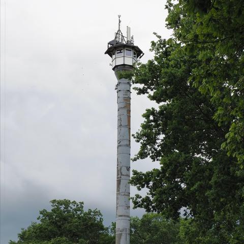 Tall observation tower with a cylindrical base and a multi-windowed cabin at the top, surrounded by trees with lush green leaves, set against a cloudy sky.