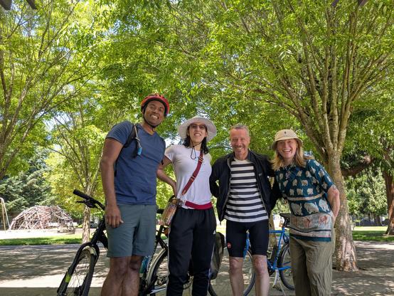 4 people standing in front of bikes with a park backdrop with lots of trees, and a jungle gym dome.   Second from the left is me in a GALDEF t-shirt, and white sun hat, 4th from the left is Carrie another Genital Autonomy Collective Board Member.