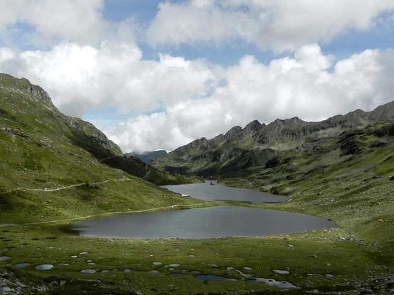 Zwei klare Gebirgsseen hintereinander inmitten von grüner Fläche und begrünten Hängen, durch die Wanderwege verlaufen. Zwischen den Seen ist ganz klein eine Hütte zu erahnen. Zum Horizont hin verlaufen rechts und links Gebirgskämme, über allem meist helle Wolkenbänder, die vor blauem Himmel Schatten in der Szenerie verteilen.