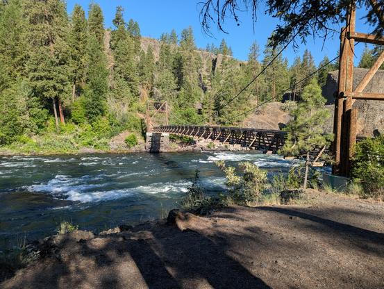 view of the Spokane River at the bowl and pitcher campground at Riverside State Park. the river is running across and there's a wooden bridge going over the river off into the trails