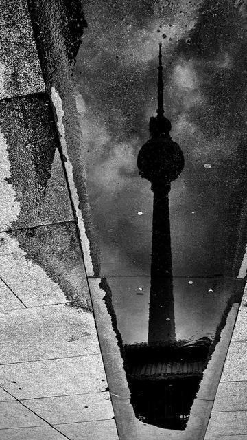 Reflection of a tower in a puddle, captured in black and white. Surrounding pavement shows texture and lines.