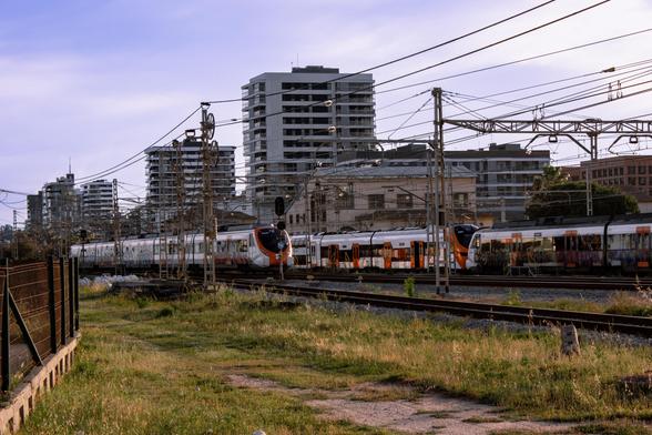 A view of the Renfe 452 from afar. There's a vandalized Civia passing by next to it. Some rather tall buildings are at the background.
