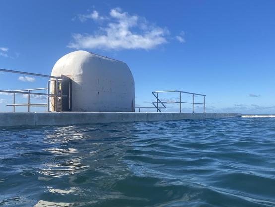 Rounded white pump house building on the northern (left) side of the pool on a sunny winter's day. Sky is pale near the horizon, gradually intensifying to mid blue. There's a thin wisp of cloud above the pump house.  The water in the baths is a deeper blue with choppy white reflections of the pump house.