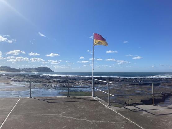 Looking towards the Bar Beach headland from the Merewether Baths on a sunny winter's day. The red and yellow surf life saving flag is flying to the right. Blue sky with small clumps of white cloud.