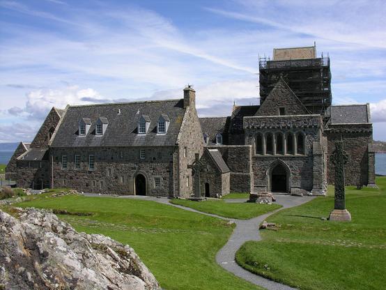 Iona Abbey. The image shows an abbey with sea in the background. The abbey church is seen end-on on the right and other buildings are to its left. There is mown grass with paths in the foreground and an outcrop of rock on the lower left of the frame. The scene is in sunlight.