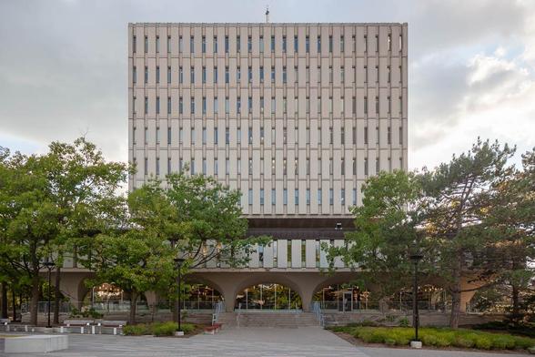 A rectangular, multi-story concrete building with vertical window slats and an elevated first floor supported by large arches, surrounded by trees and a paved plaza.