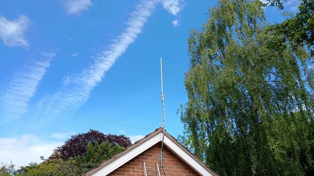 A vertical antenna attached to a short aluminium mast affixed to the apex of a brick garage wall. There is a tall tree to the right of the antenna. The sky is blue, with a few white clouds.