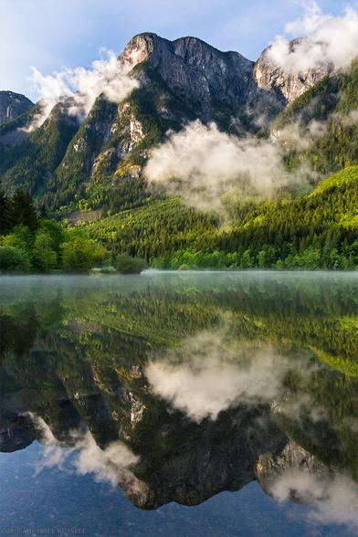 The still water of a lake reflect nearly perfectly a mountain peak on the other side of the lake, along with the green foliage and clearing clouds.