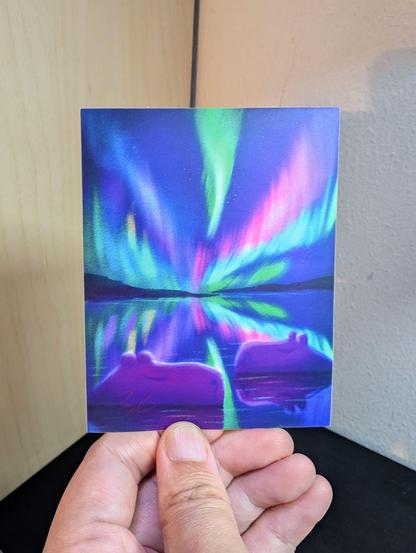 Photo of a left hand holding a rectangle sticker. The sticker is a matte painting of two capybaras under neon northern lights.