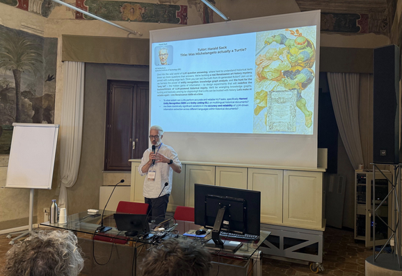 Harald Sack, co-director of ISWS, is presenting his research task, standing in front of a screen, where besides the title (Was MIchelangelo really a Turtle?" an AI-generated illustration is showing MIchelangelo (the artist) in fight with Michelangelo, the Teenage Mutant Ninja Turtle, is depicted.