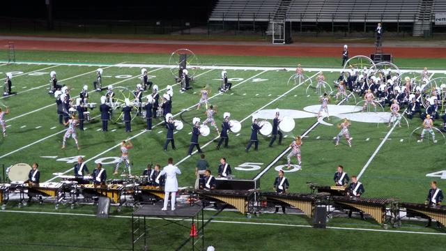 A lively outdoor performance featuring a marching band and color guard on a football field at night. Musicians in uniforms play various instruments, while performers in vibrant costumes showcase choreographed movements. Elements like large drums and marching formations create an energetic atmosphere.