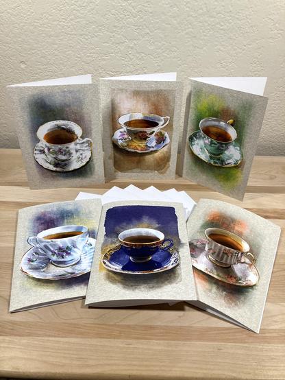 Photograph of six illustrated greeting cards featuring different teacups, arranged on a wooden plank with white envelopes.