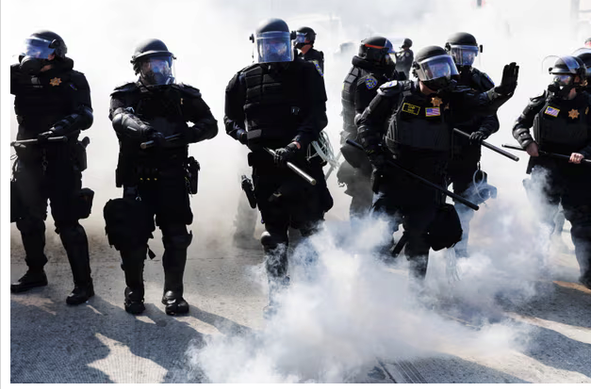 Photo: Men in riot gear and gas masks advance toward protestors, caption California highway patrol using smoke grenades to advance and push protestors off the 101 freeway during a demonstration following federal immigration operations in Los Angeles. Photograph: Blake Fagan/AFP/Getty Images