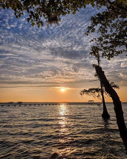 Colorful sunset as golden shades of yellow and orange stretch across the horizon, beneath big blue skies with layered rows of white clouds, and above gentle waves of a vast waterway reflecting a bright beam of glowing light across the surface.