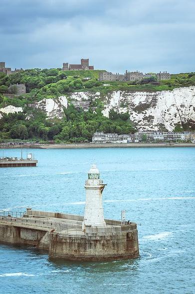 A lighthouse stands prominently at the edge of a pier, overlooking calm blue waters. In the background, majestic white cliffs and lush greenery provide a striking contrast, with a historic castle and church perched atop the hill. Photography by Debra Martz