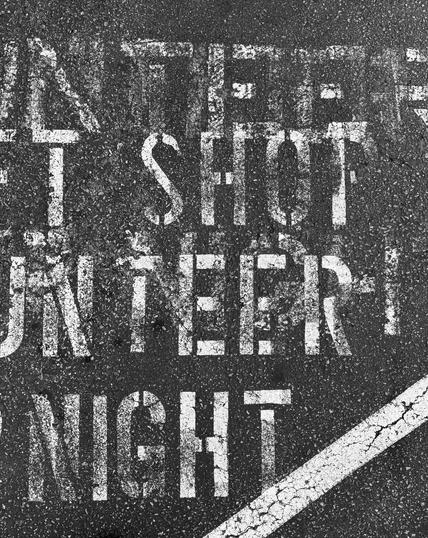 The image shows a close-up of a textured asphalt surface with white stencil text and a white line. The text is partially visible, with layered words and appears to read "ANTIFIRE SHOT UNTEER NIGHT”, although the actual words stenciled on the parking lot say something about parking for the volunteer shop only, at all times, day and night. The letters are large, bold, and somewhat faded, indicating weathering or wear. The white line is a diagonal crack, suggesting age and exposure to the elements. The overall color scheme is monochromatic, with the stark contrast between the white text and line against the dark asphalt. The image captures the gritty texture of the asphalt, with small stones and debris visible in the background.