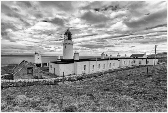A black and white image of a lighthouse surrounded by buildings, set against a cloudy sky and overlooking the sea. The landscape features grassy terrain and stone walls.