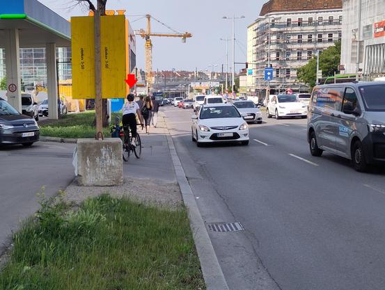 Foto der dreispurigen Richtungsfahrbahn, eine Person radelt am Gehsteig gegen die Fahrrichtung. Eine Fahrbahn, die nach Radweg aussieht, endet vor einer Tankstelle.