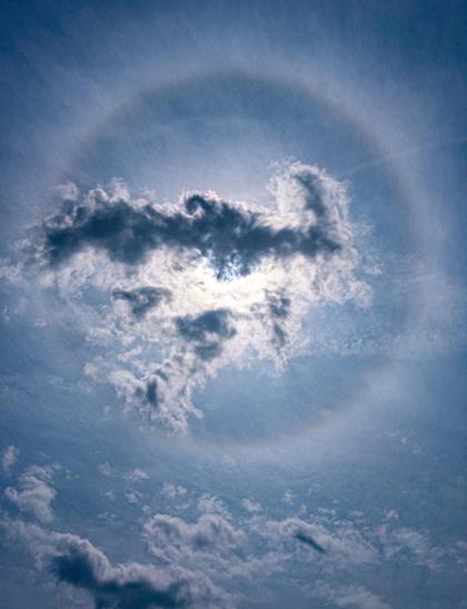 The sun shining partly through a cloud with cirrus in the blue sky around. There is a 22 degree halo encircling the sun.