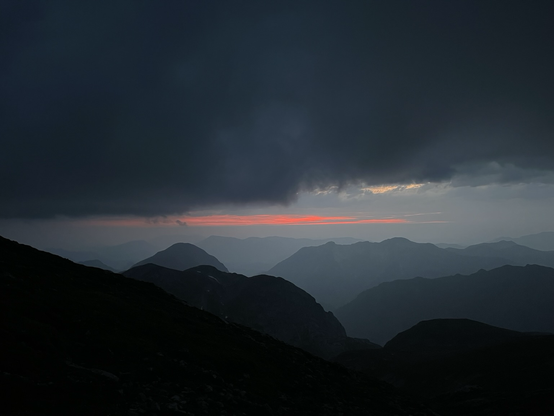 Very dark clouds above, with a small patch of orange clouds in the middle, low contrast dark outlines of a lot of mountains below.