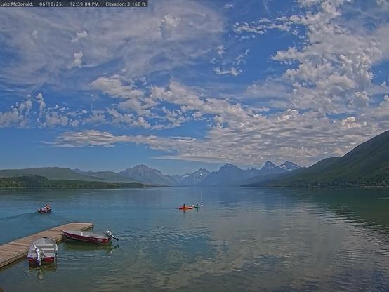 Park visitors enjoy paddling on Lake McDonald in Glacier National Park today as blue sky and interesting white clouds are reflected on the lake’s surface. A dock with red, white, and silver boats can be seen on the left. Green trees and distant snow-covered mountains provide a backdrop for the lake. Thanks to the National Park Service and the Glacier National Park Conservancy for these beautiful views.