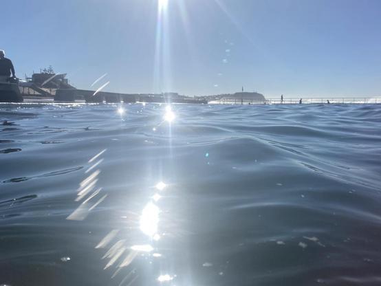 Sun glitter on the dark blue water. Taken while swimming in the main pool at the ocean baths looking northwards to the Bar Beach headland. The white spray of the breaking waves at nearby Merewether Beach is visible above the rim of the pool.