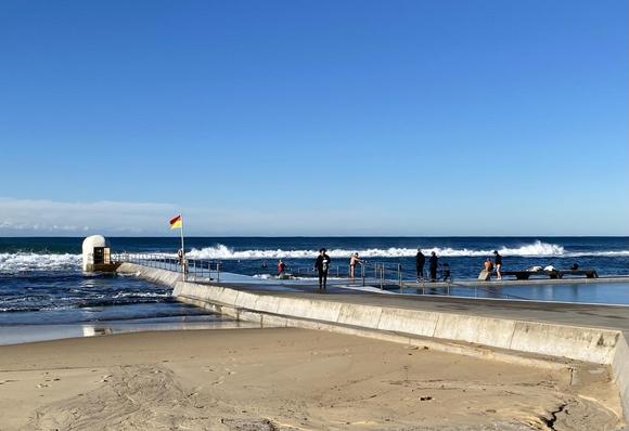 Looking over the northern concourse of the ocean baths and out to sea. The white rounded pump-house marks the north east corner on the far left with the red and yellow surf life saving flag flying closer to the viewer. A few swimmers in the pool, some getting in or out. A few in winter clothes standing or walking nearby. White waves breaking along the back wall. Lots of blues - sky and water.