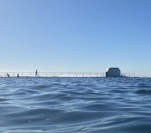 Looking across the dark blue water towards the northern side of the main pool. The white rounded pump house building is on the right. There are three figures in silhouette on the left: a swimmer leaving the pool via the ramp, one entering, and a walker striding along the path above. Lots of clear blue sky.