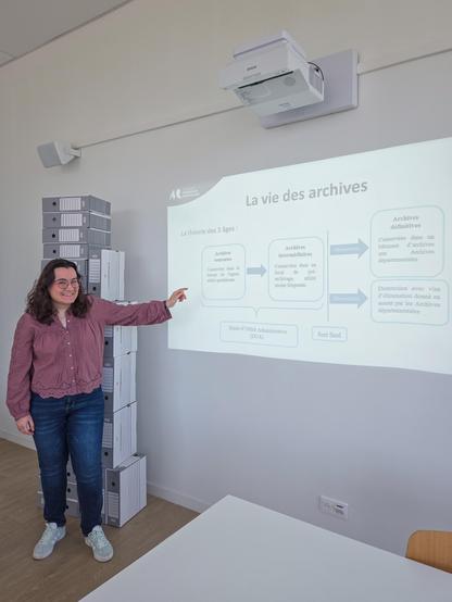 Modestine Lesquoy, présentant la vie des archives - pile de boîtes à archives derrière elle