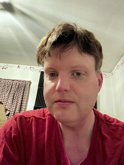 A rare selfie. I’m wearing a red t-shirt and am posing nonchalantly. My hair is dirty blonde and my eyes are a greyish blue. Soundproofing panels adorn the walls of my office/home studio in the background.