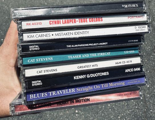 A hand holding a stack of CDs, including titles by Cyndi Lauper, Kim Carnes, Cat Stevens, Kenny G, Blues Traveler, and Amy Grant, on a pavement surface.
