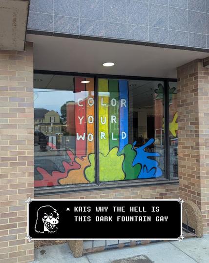 The image shows a storefront with a large window display featuring a colorful design. The window is divided into vertical sections, each filled with a different color: red, orange, yellow, green, and blue. The colors are vibrant and create a rainbow effect. The text "COLOR YOUR WORLD" is written in white capital letters, with each word on a separate colored background. Below the window, there is a black text box with a white pixel art face of the character Suzie from Deltarune on the left. The text box reads "* KRIS WHY THE HELL IS THIS DARK FOUNTAIN GAY"