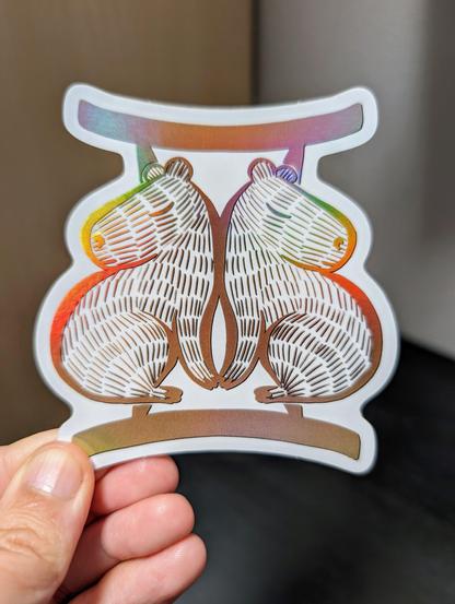Photo of a left hand holding a diecut sticker. The sticker is a holographic drawing of a capybara Gemini zodiac sign.