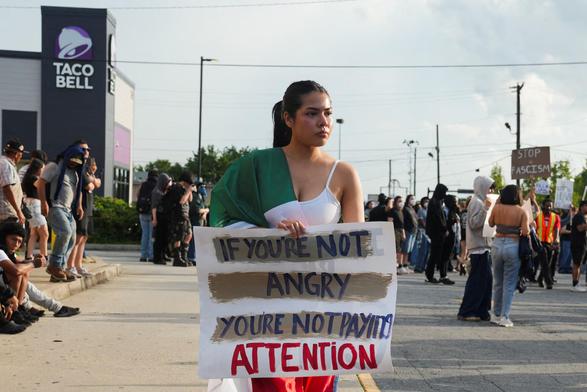 A young woman wrapped in a green, white & red flag carries a sign which reads:
IF YOU'RE NOT ANGRY YOU'RE NOT PAYING ATTENTION