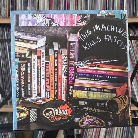 Eine künstlerische Auslage mit Büchern, darunter Titel wie "Let It Bleed" und "American Hardcore", neben dekorativen Elementen und dem auffälligen Satz "This Machine Kills Fascists" im Kreidestil. Im Hintergrund sind Schallplatten in Regalen zu sehen.