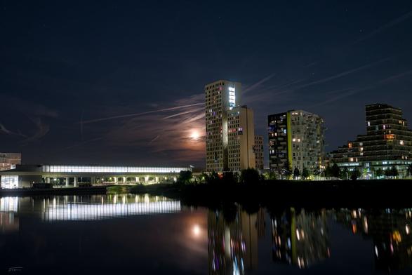 Das Foto zeigt eine nächtliche Stadtansicht mit modernen Hochhäusern am Ufer eines ruhigen Gewässers. Im Hintergrund ist der Mond zu sehen, der teilweise von Wolken und Kondensstreifen verdeckt wird und einen sanften Lichtschein verbreitet. Die Gebäude sind beleuchtet, und viele Fenster strahlen in unterschiedlichen Farben. Die fast weißen Lichter der U-Bahn-Station Seestadt spiegeln sich klar auf der glatten Oberfläche des Sees wider, was dem Bild eine stimmungsvolle und ruhige Atmosphäre verleiht