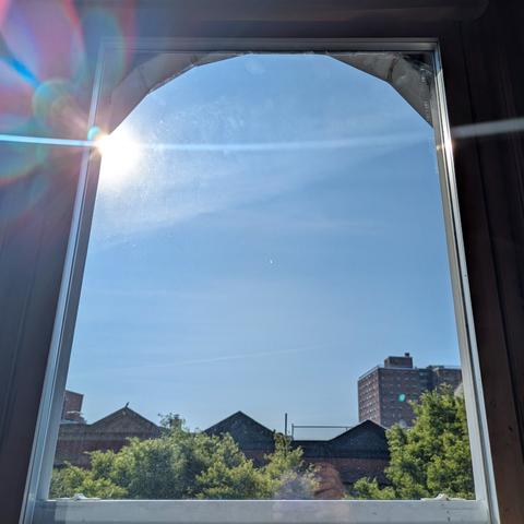 Looking through an arched window three hours after sunrise the sun is above the buildings, making a dramatic lens flare that bisects the frame horizontally with a curved white line and multi-colored petals above. The sky is light blue with thin white clouds. Pointed roofs of Harlem brownstones are silhouetted across the street, and a taller apartment building can be seen in the distance. The green tops of two trees are on the bottom and right.
