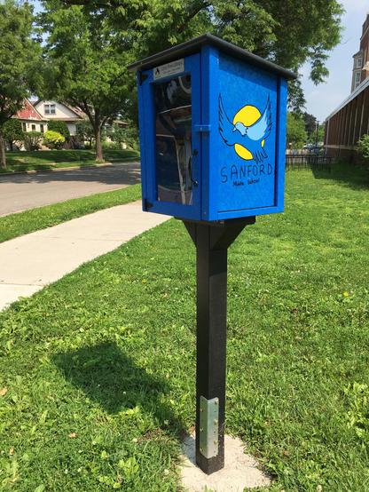 A Little Free Library on the corner of the ground of Maria Sanford Middle School in the Longfellow neighborhood, near the river in south Minneapolis. It's been painted blue and black and the logo consists of a bird flying in front of the Sun. "Sanford Middle School." Metal angle pieces have repaired the wooden post which supports it.