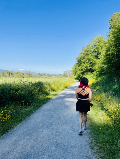 Crissy wearing black hat, top, skirt walking towards a path. Surrounded by green flowering plants on left, trees on right. You can see Vancouver in the distant skyline