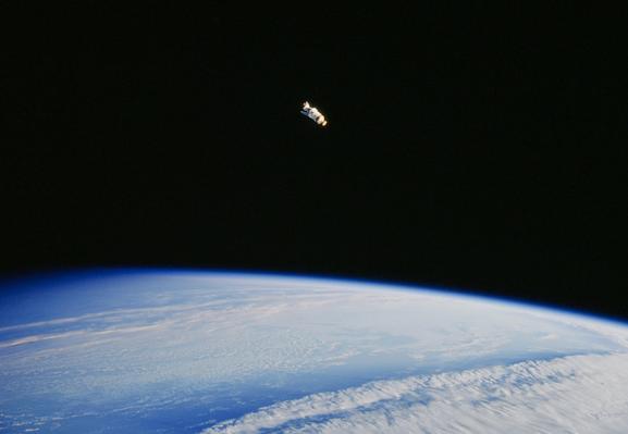 An image of dark space with a cloudy white and blue Earth at the bottom and a white and yellow spacecraft in the middle towards the top.