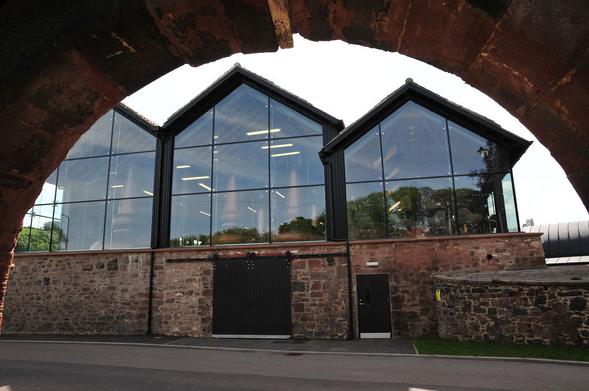 Lindores Abbey Distillery. The image shows a view out from under a stone arch. On the opposite side of a road and largely framed by the arch is a glass-fronted distillery building. The lower part is in stone, and the upper part, comprising three gable ends side by side is glass. The stills can be seen inside.