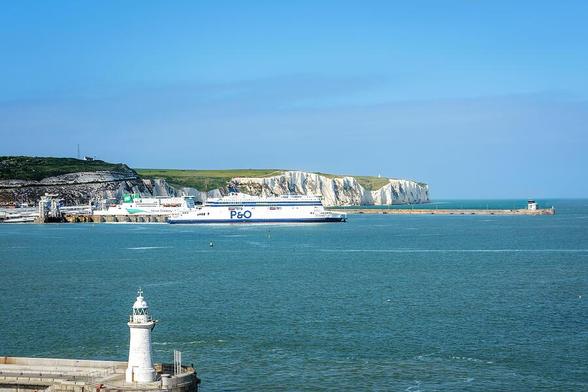 A ferry is docked and one is departing the port, with the iconic white cliffs of Dover in the background and a lighthouse in the foreground. The sea is calm under a clear blue sky. Photography by Debra Martz