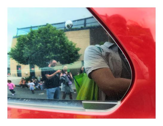 An outdoor city scene is reflected in the window of a parked red car. The reflection starts from left frame and takes up about 2/3rds of the image, bordered by the window’s curved diagonal edges. The car door’s glossy red surface serves as a frame at bottom and right. The reflection shows adults standing in the street while checking their phones, small children running around, a sidewalk tree, and a low-rise apartment building. I can be seen in the foreground of the reflection —  my arm and green tote bag appear in the window panel, while my face and my hand holding the iPhone are blurred by the red doorframe.