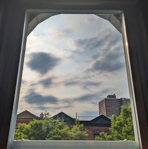 Looking through an arched window two and a half hours after sunrise the sky is full of windswept white and grey-blue clouds. Pointed roofs of Harlem brownstones are silhouetted across the street, and a taller apartment building can be seen in the distance. The green tops of two trees are on the bottom and right.