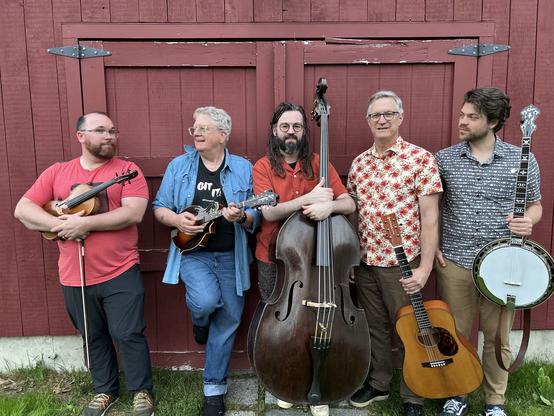 A group of five musicians stands in front of a red barn. Each holds a musical instrument: a violin, mandolin, double bass, acoustic guitar, and banjo. They are dressed casually, with varying patterns and colors. The setting is outdoors